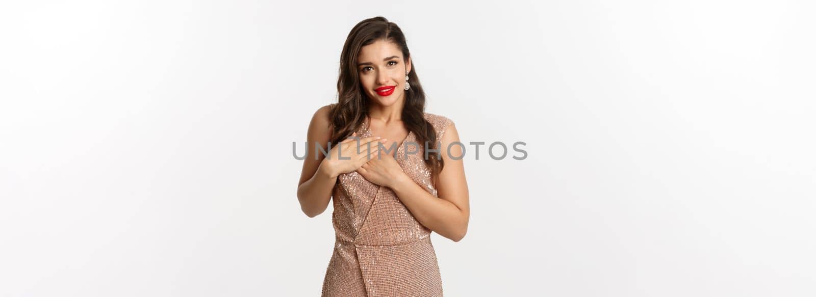 Image of beautiful young woman staring grateful at camera, holding hands on heart touched and thankful, receiving gift, standing in evening dress at party, white background.