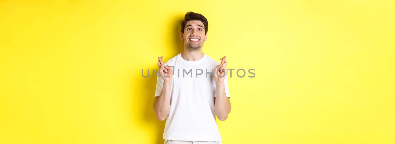 Nervous and hopeful man praying to god, making wish with fingers crossed, panicking and standing over yellow background by Benzoix