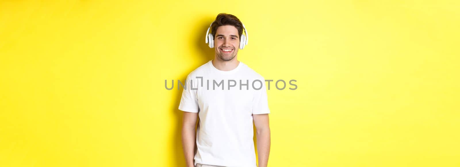 Young handsome man listening music in headphones, wearing earphones and smiling, standing over yellow background.