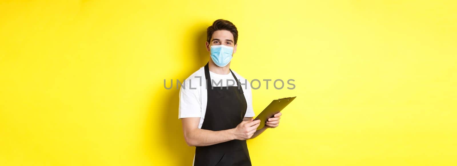 Concept of covid-19, small business and quarantine. Young male seller in medical mask and black apron taking orders, holding clipboard, standing over yellow background.