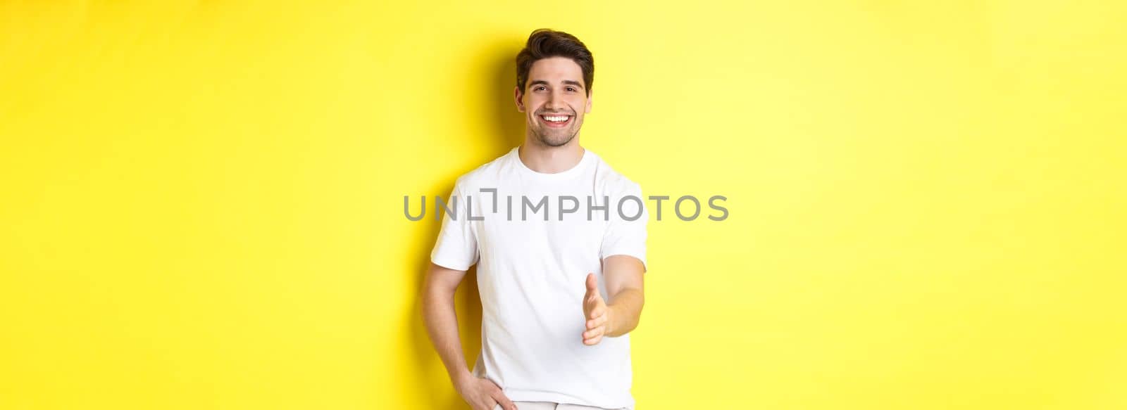 Handsome and confident man extending hand for handshake, greeting you, saying hello, standing in white t-shirt over yellow background by Benzoix