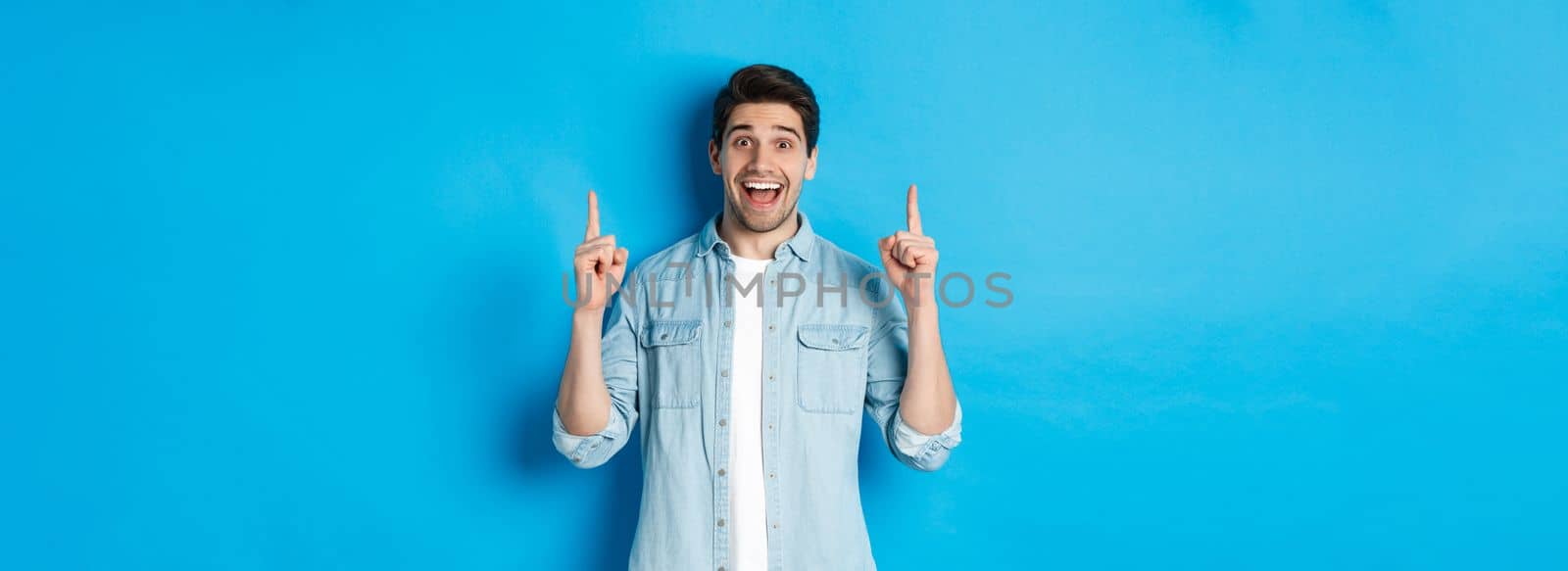 Portrait of happy 25s guy with beard, pointing fingers up and smiling, showing advertisement, standing against blue background.