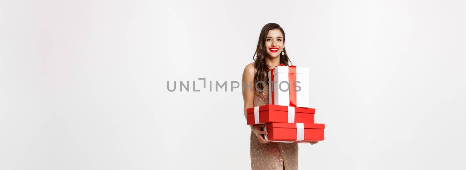Christmas party and celebration concept. Full-length of stylish woman with red lips, glamour dress, holding gifts and smiling happy, standing over white background.
