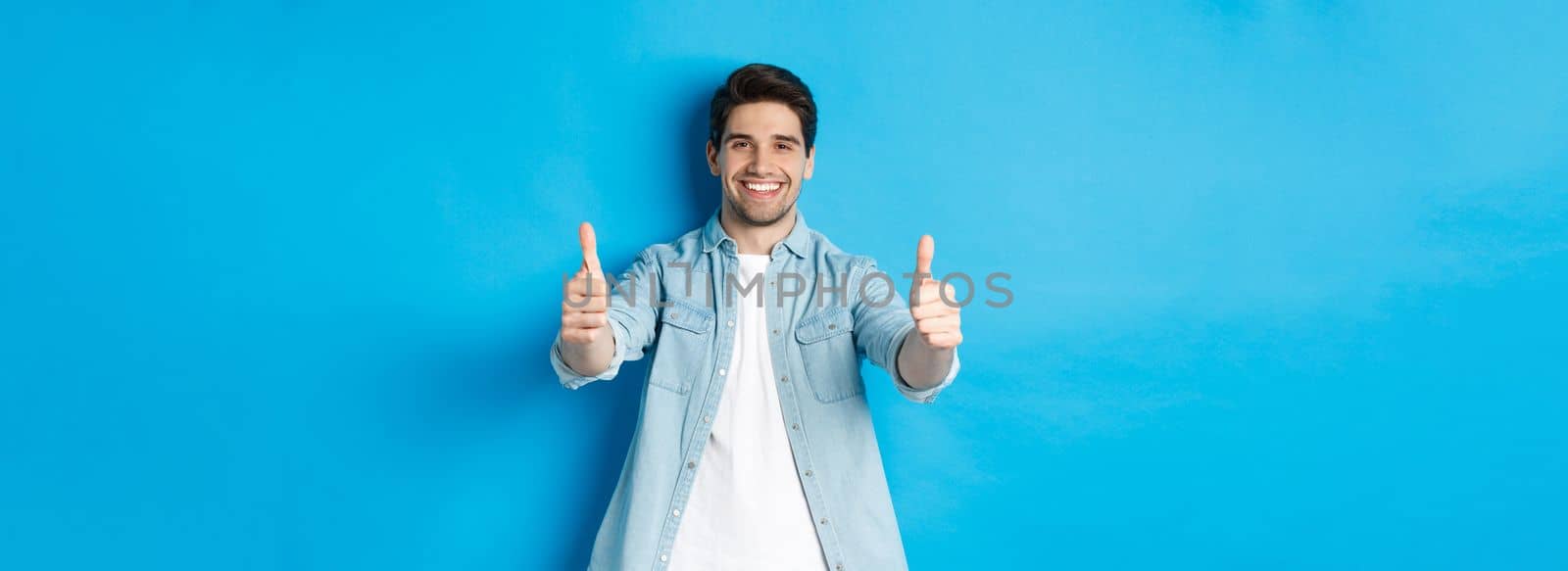 Smiling confident man showing thumbs up, guarantee quality, approving something good, standing against blue background by Benzoix