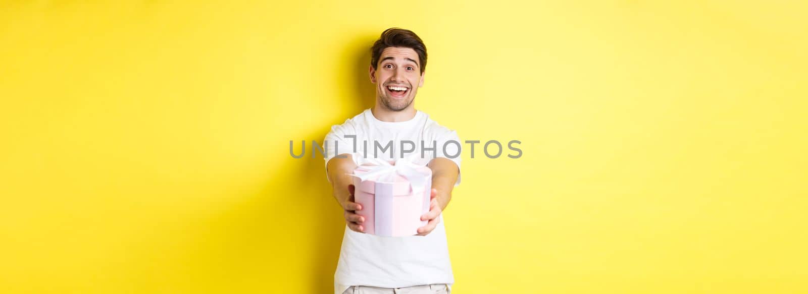 Concept of holidays and celebration. Smiling man giving you gift, congratulating, standing over yellow background with present by Benzoix