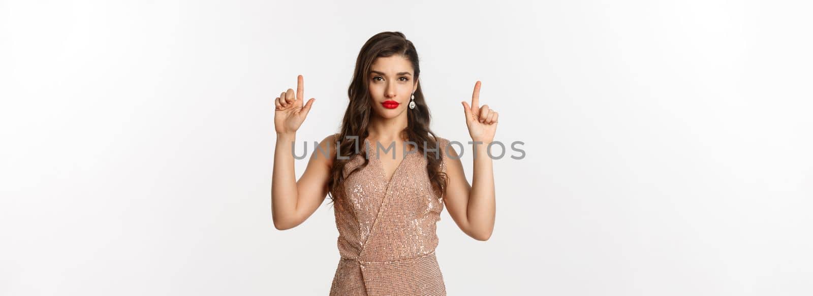 New Year, christmas and celebration concept. Elegant young woman with red lipstick, wearing party dress and looking sassy, pointing fingers up at logo, white background.