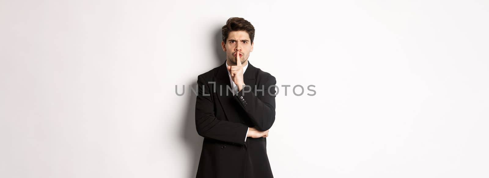 Portrait of angry boss in suit shushing at you, telling to be quiet, showing taboo hush sign and frowning, standing over white background.