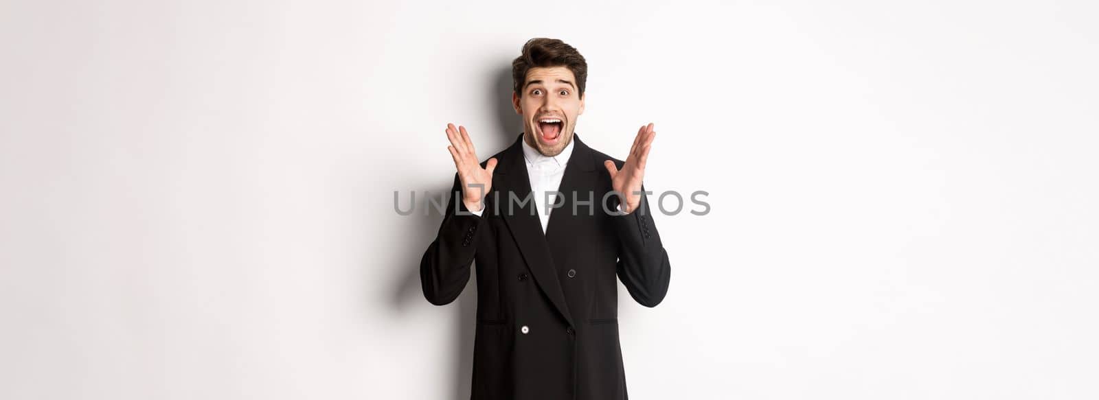 Portrait of surprised handsome businessman in suit, reacting to holidays promo, looking amazed at camera, standing over white background by Benzoix