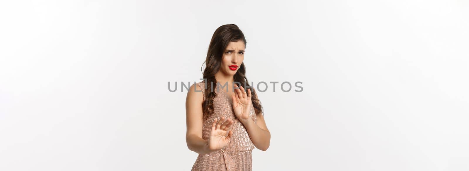 Concept of celebration, holidays and party. Woman looking displeased and rejecting something, wearing glamour dress, showing stop gesture, asking stay away, white background.