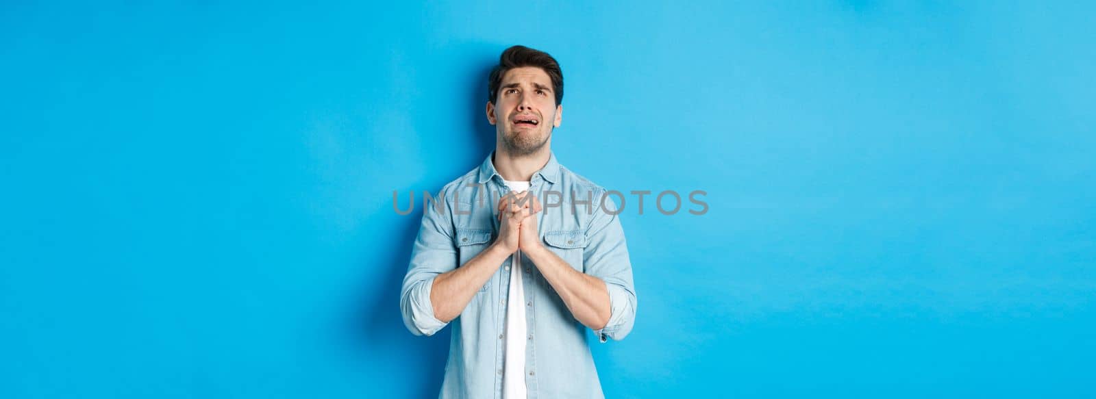 Desperate man begging and crying for help, pleading to god, looking up and holding hands in pray, standing against blue background.
