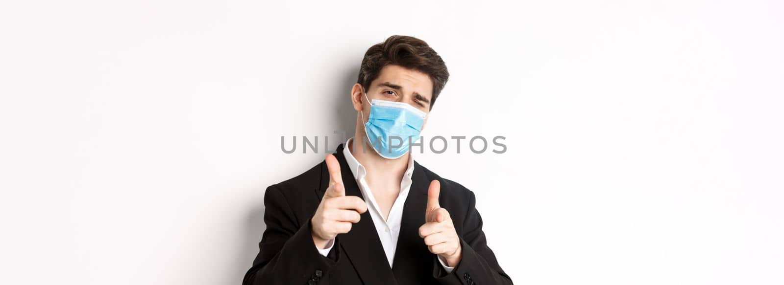 Concept of covid-19, business and social distancing. Close-up of sassy man in trendy suit and medical mask, pointing fingers at camera, standing against white background by Benzoix
