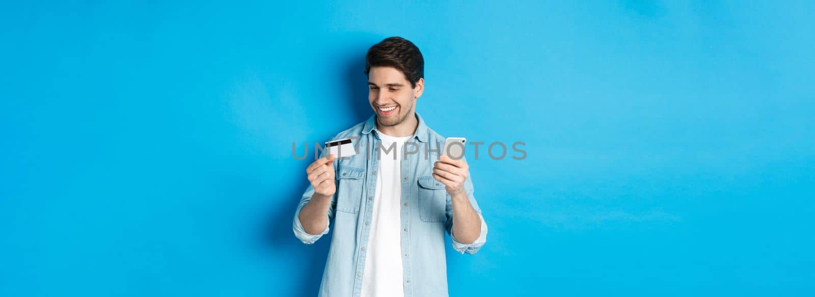 Handsome happy man paying for something online, holding credit card and mobile phone, purchase in internet, standing over blue background.