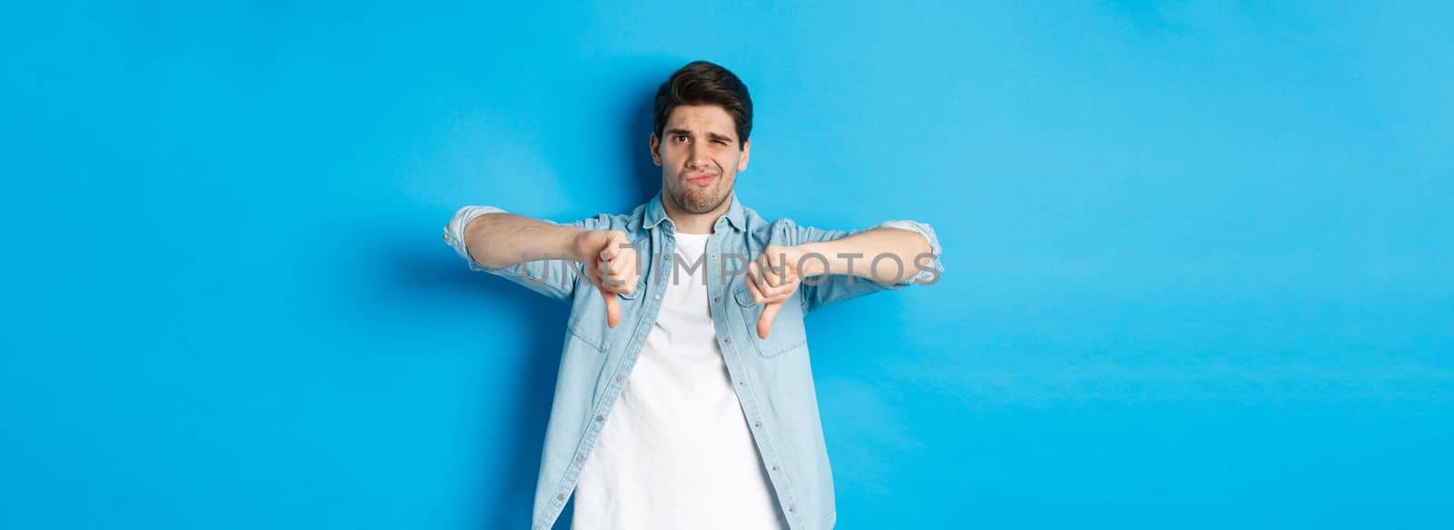 Skeptical and disappointed 25 years old man in casual outfit dislike something bad, showing thumbs-down and grimacing displeased, standing against blue background.