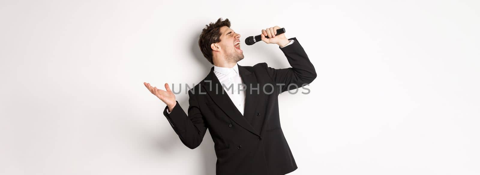 Portrait of handsome man singing a song with passion, standing in black suit, holding microphone and performning, posing over white background by Benzoix