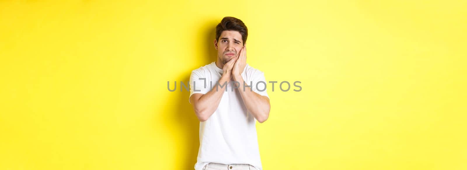 Sad guy complaining on toothache, touching swallen cheek and looking upset, standing over yellow background. Copy space