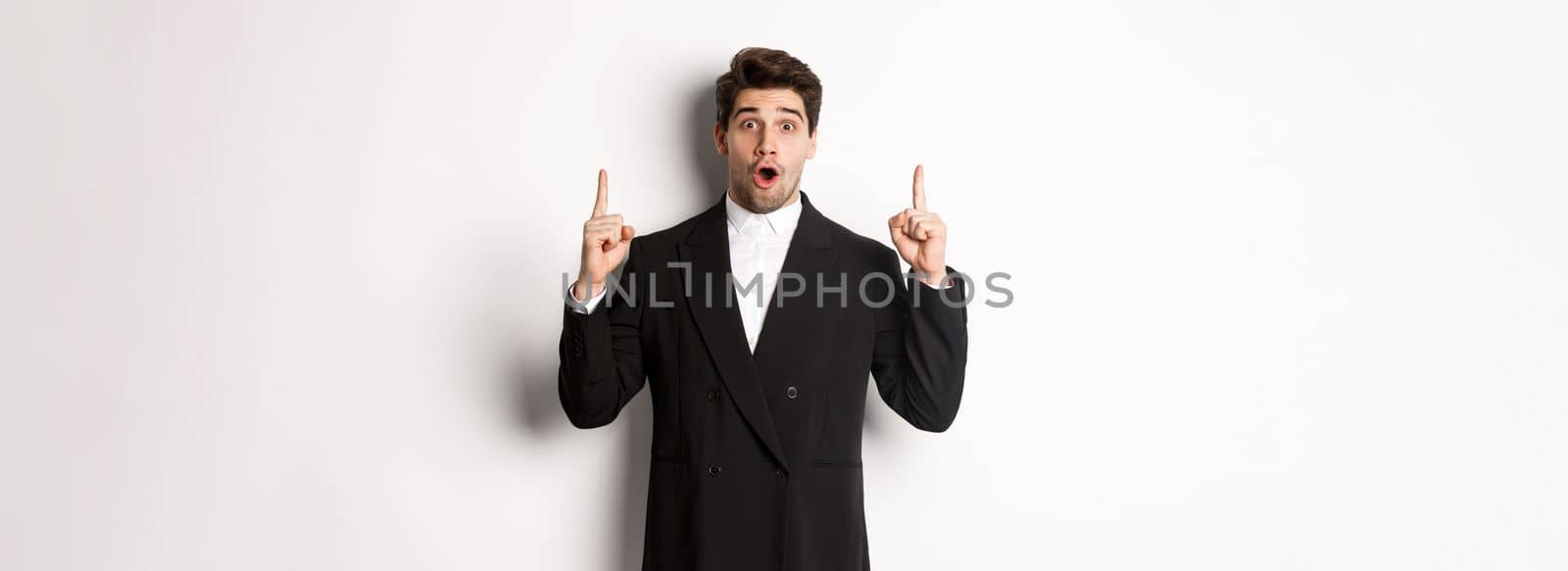 Portrait of surprised handsome businessman in black suit, saying wow and looking amazed, pointing fingers up at copy space, standing over white background.