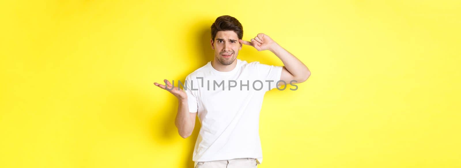 Confused and angry man pointing at head, scolding person for acting stupid, show crazy sign, standing over yellow background by Benzoix