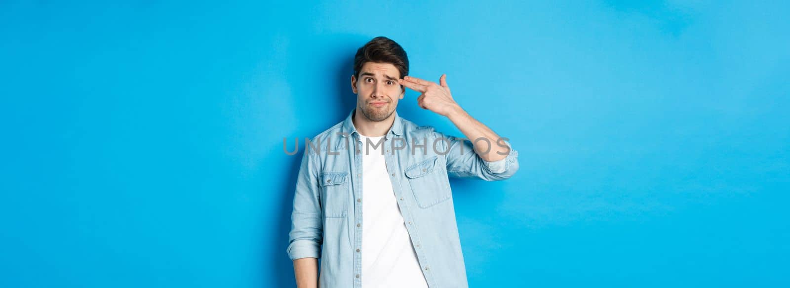 Tired and annoyed adult man making finger gun gesture near head, shooting himself from boredom, standing against blue background by Benzoix