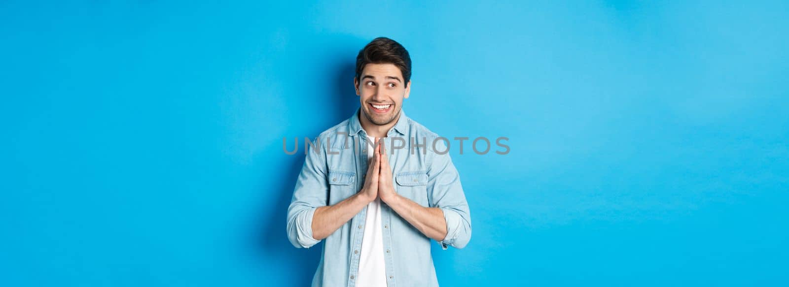 Hopeful smiling man looking left, holding hands in pray, begging for something or pleading, standing against blue background.
