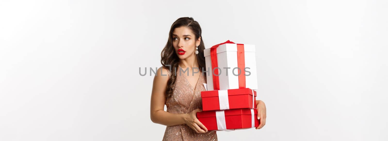New Year, Christmas and celebration concept. Attractive young woman in elegant dress, looking away, carry gifts, standing over white background by Benzoix