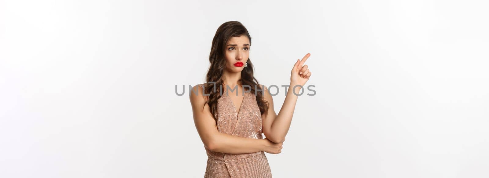 Christmas, holidays and celebration concept. Skeptical and displeased woman in stylish party dress, pointing finger left and starring at banner unamused, white background.