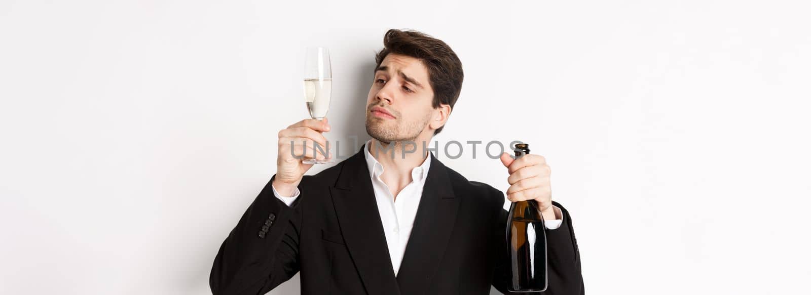 Close-up of attractive man in trendy suit, tasting champagne, looking at glass, standing against white background.