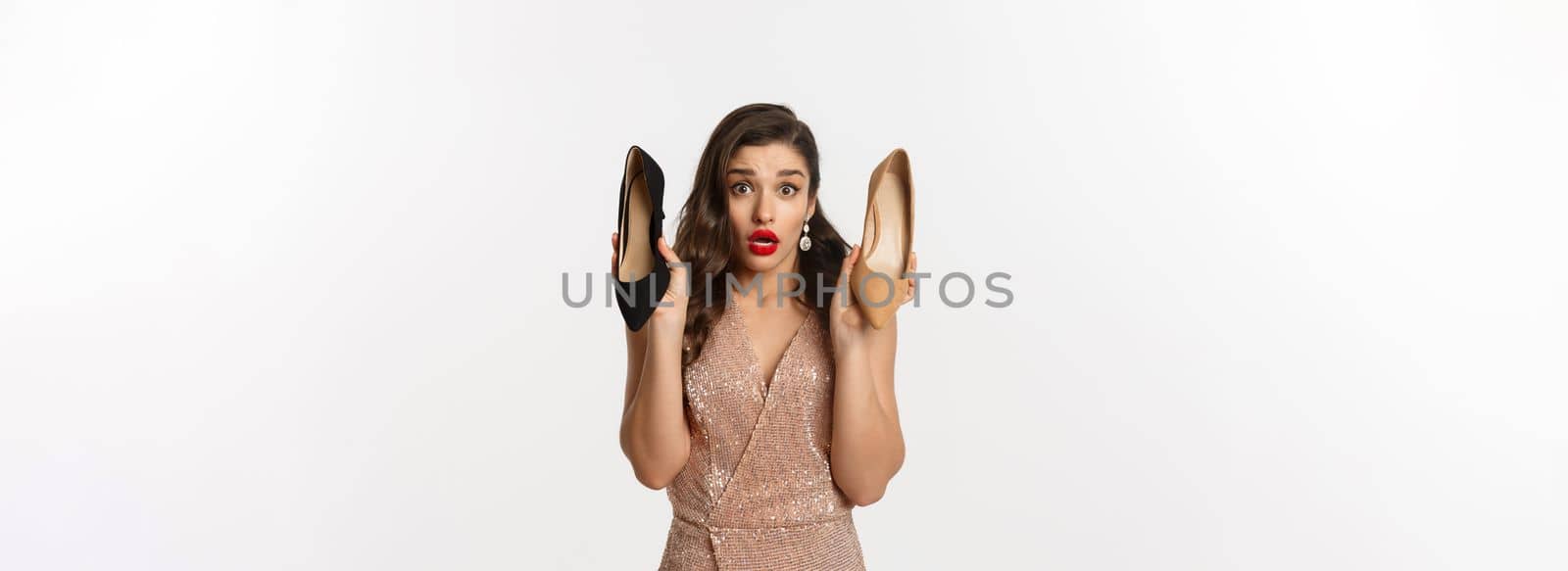 Party and celebration concept. Indecisive girlfriend showing two different heels, asking opinion, help with choice while picking outfit, standing over white background.