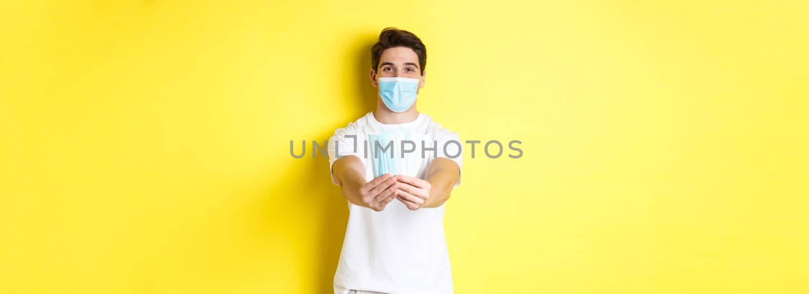 Concept of covid-19, quarantine and preventive measures. Young caucasian man giving medical masks for you, standing against yellow background by Benzoix