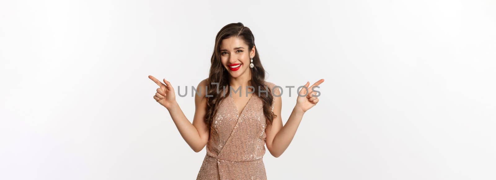 Party. Beautiful woman in elegant dress, with red lips, pointing fingers sideways, showing left and right promo offers, smiling and looking at camera, white background.