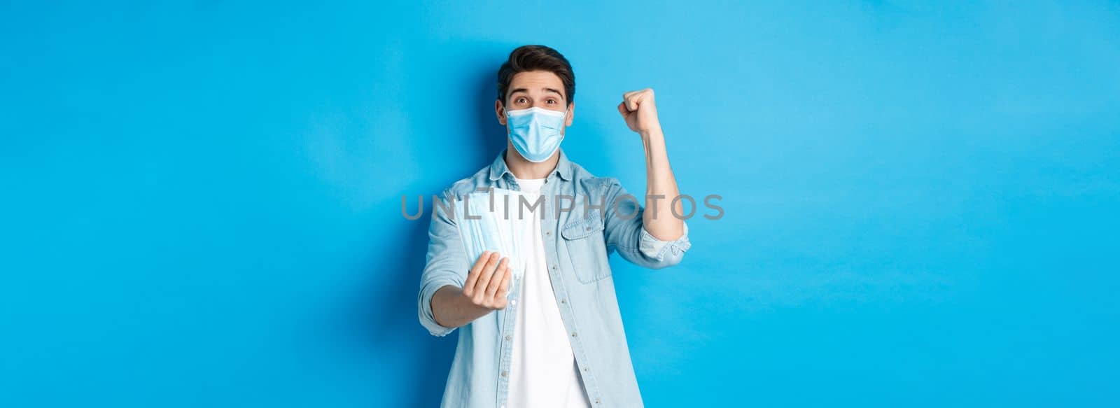 Concept of coronavirus, quarantine and social distancing. Cheerful man showing medical masks and making fist pump, celebrating or triumphing, standing against blue background.