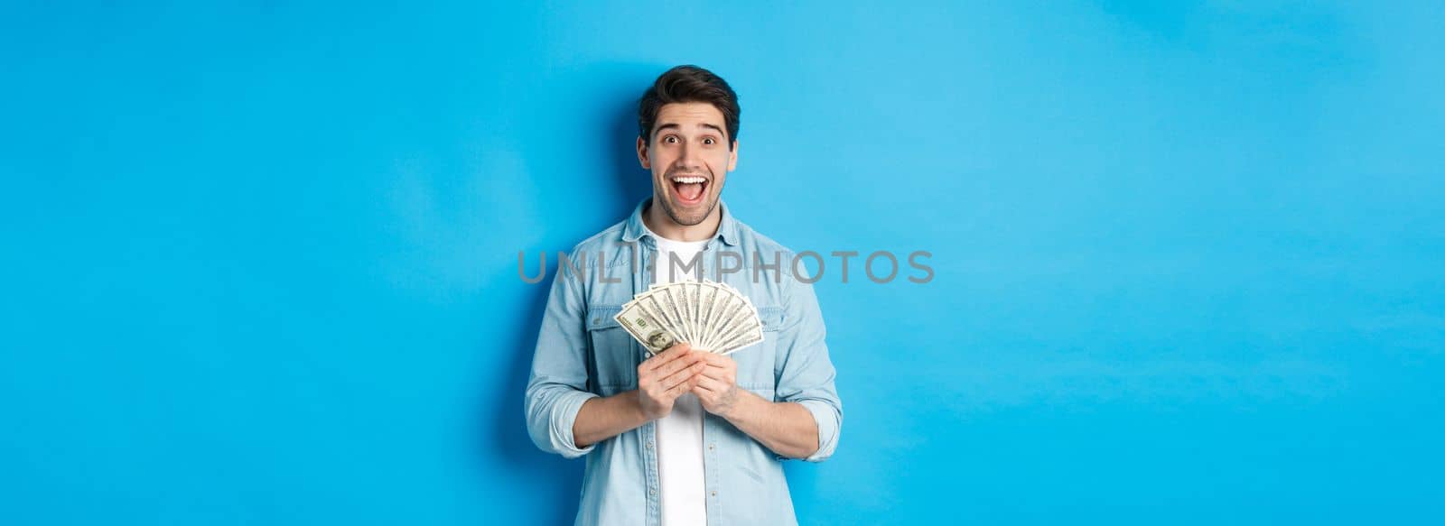 Excited and surprised attractive man, holding money prize and smiling amazed, standing over blue background. Copy space