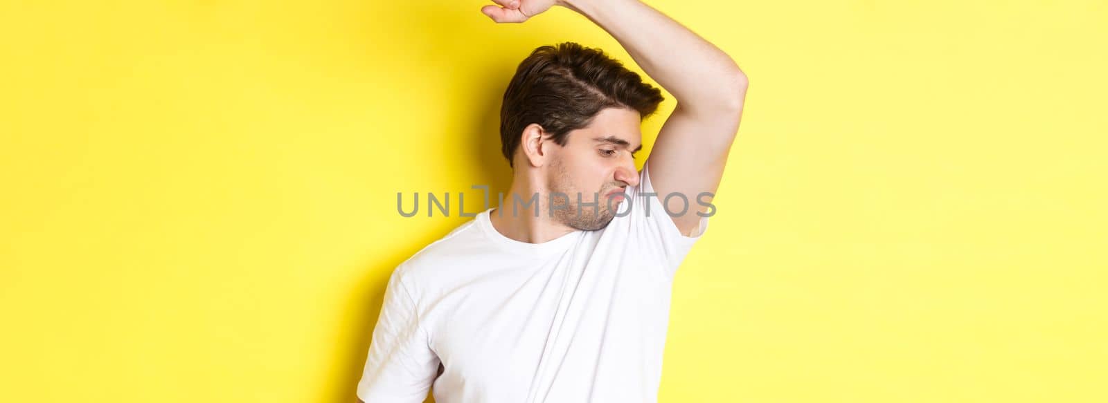 Man in sweat smelling his armpit, standing in white t-shirt and grimacing from stinky clothes.