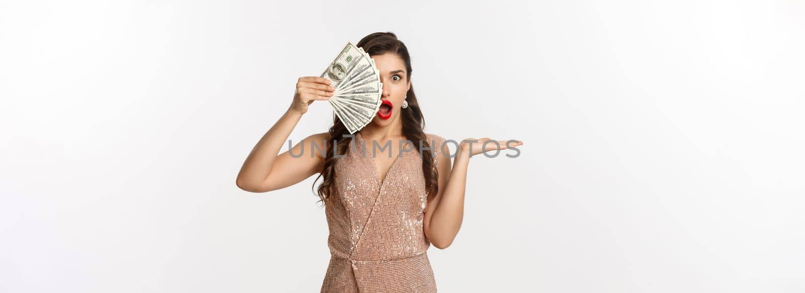 Shopping concept. Shocked woman holding money near face, wearing stylish dress, showing dollars with amazed expression, standing over white background.