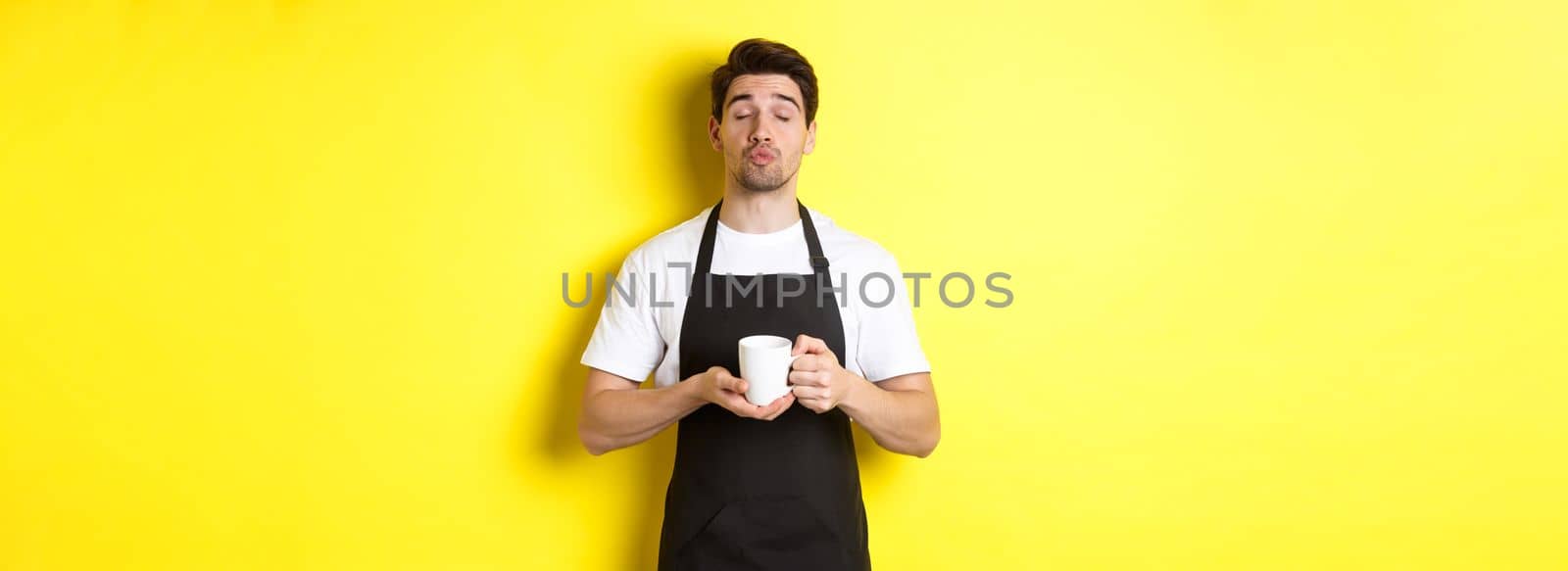 Man in black apron bringing cup of coffee and waiting for kiss, standing over yellow background.