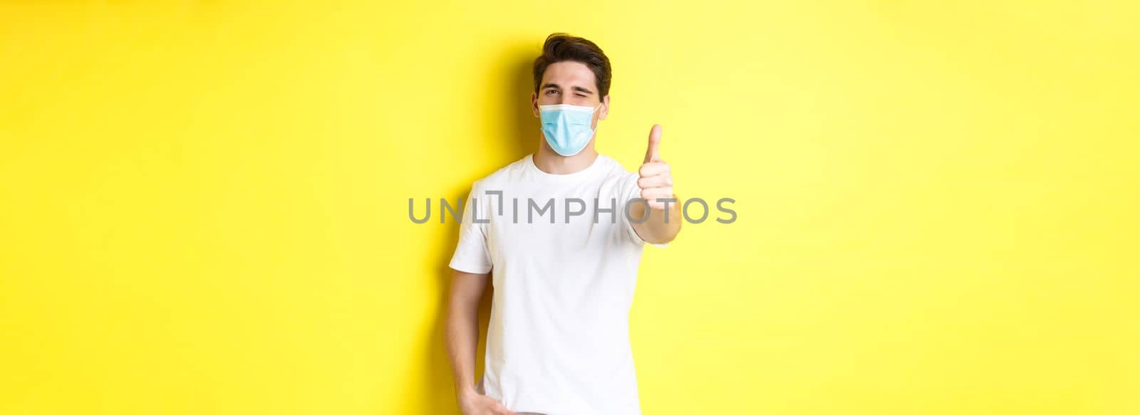 Concept of coronavirus, pandemic and social distancing. Confident young man in medical mask showing thumbs up and winking, yellow background.