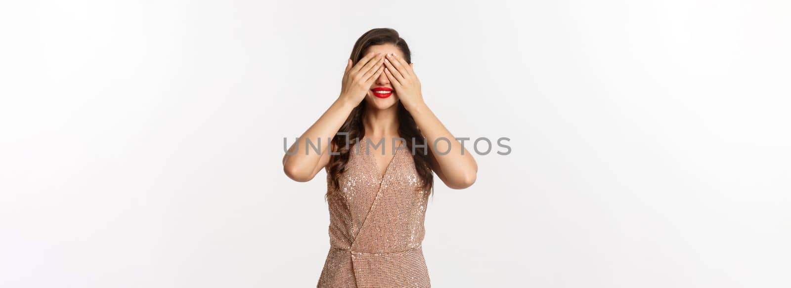 Concept of celebration, holidays and party. Attractive woman in glamour dress waiting for christmas gift, cover eyes with hands and smiling, anticipating surprise, white background.