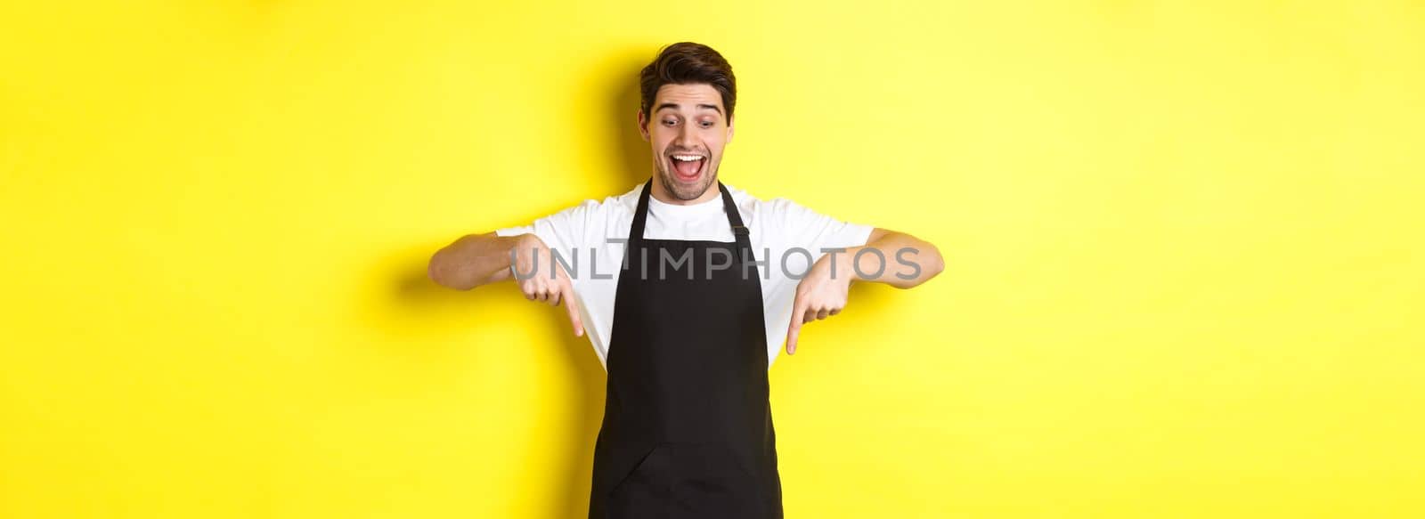 Excited waiter in black apron pointing fingers down, checking out promo offer, standing over yellow background.