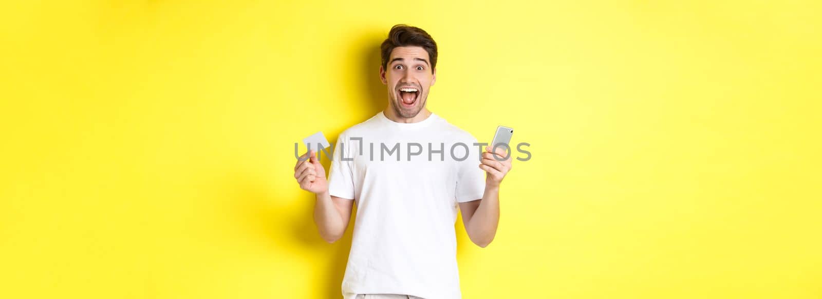 Surprised guy holding smartphone and credit card, online shopping on black friday, standing over yellow background by Benzoix