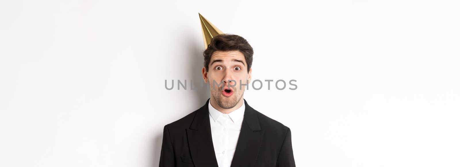 Close-up of attractive man in party hat and trendy suit, looking surprised, celebrating new year, standing against white background by Benzoix