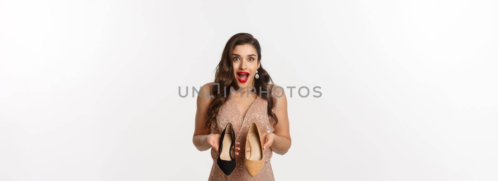 Party and celebration concept. Excited woman holding two types of heels and looking amazed, picking outfit for Christmas, standing in elegant dress over white background.