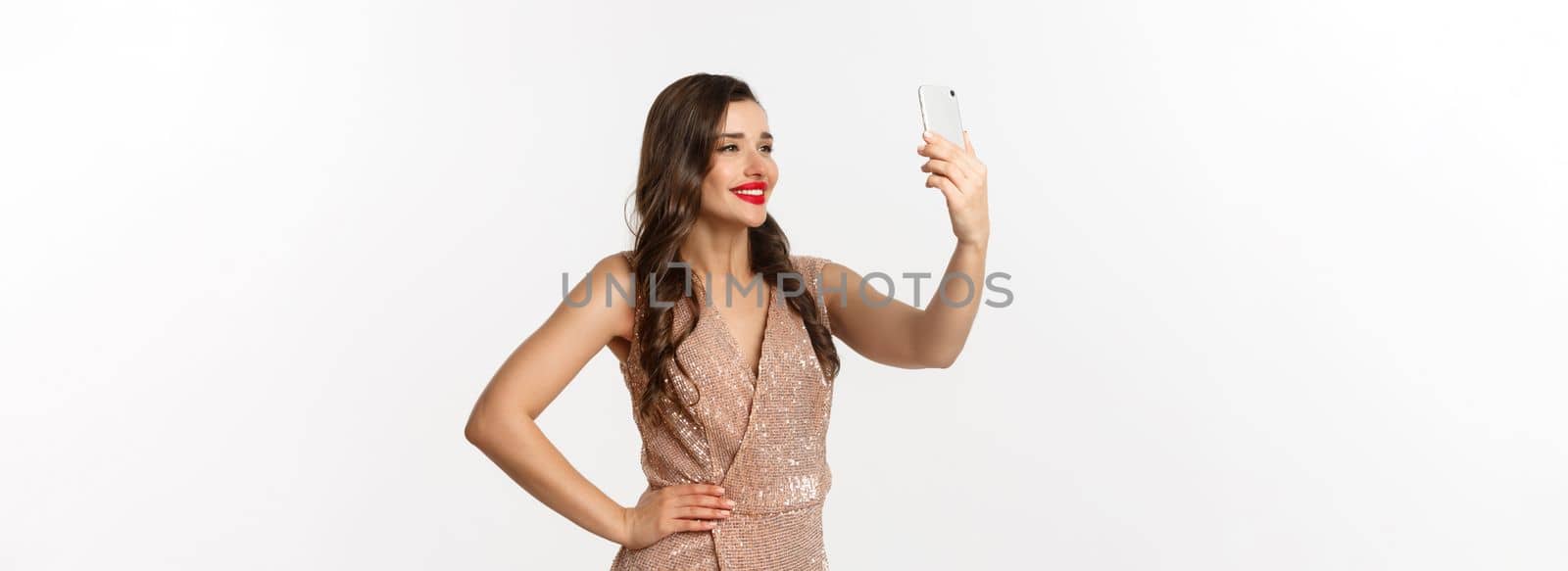 Beautiful woman with makeup and hairstyle, wearing glamour dress, taking selfie on smartphone, celebrating christmas party, white background.