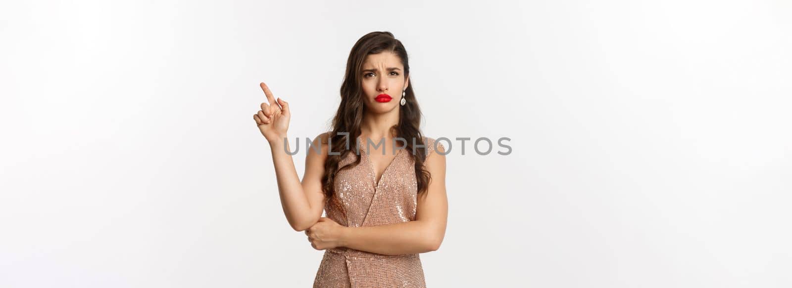 Confused glamour woman in stylish dress at new year party, pointing finger right and looking clueless, standing over white background.