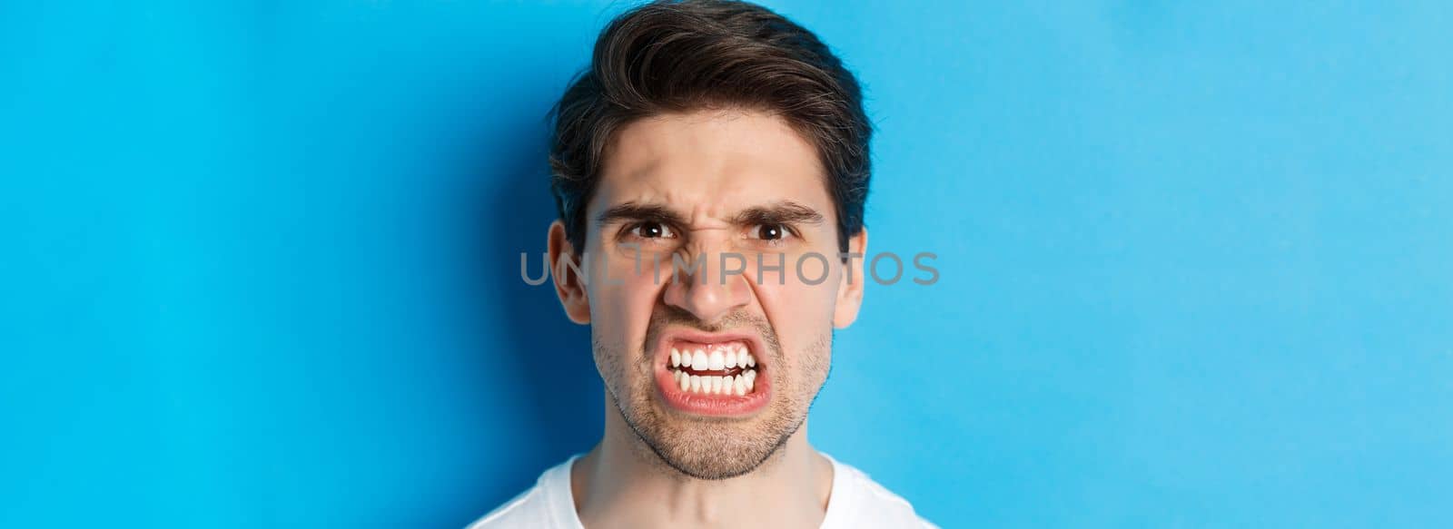 Headshot of angry caucasian man looking with scorn and dismay, being mad at person, standing against blue background.