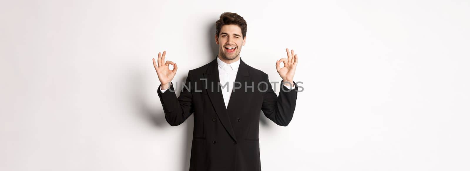 Concept of new year party, celebration and lifestyle. Portrait of successful handsome businessman in suit, winking and showing okay signs, recommending something good, white background.