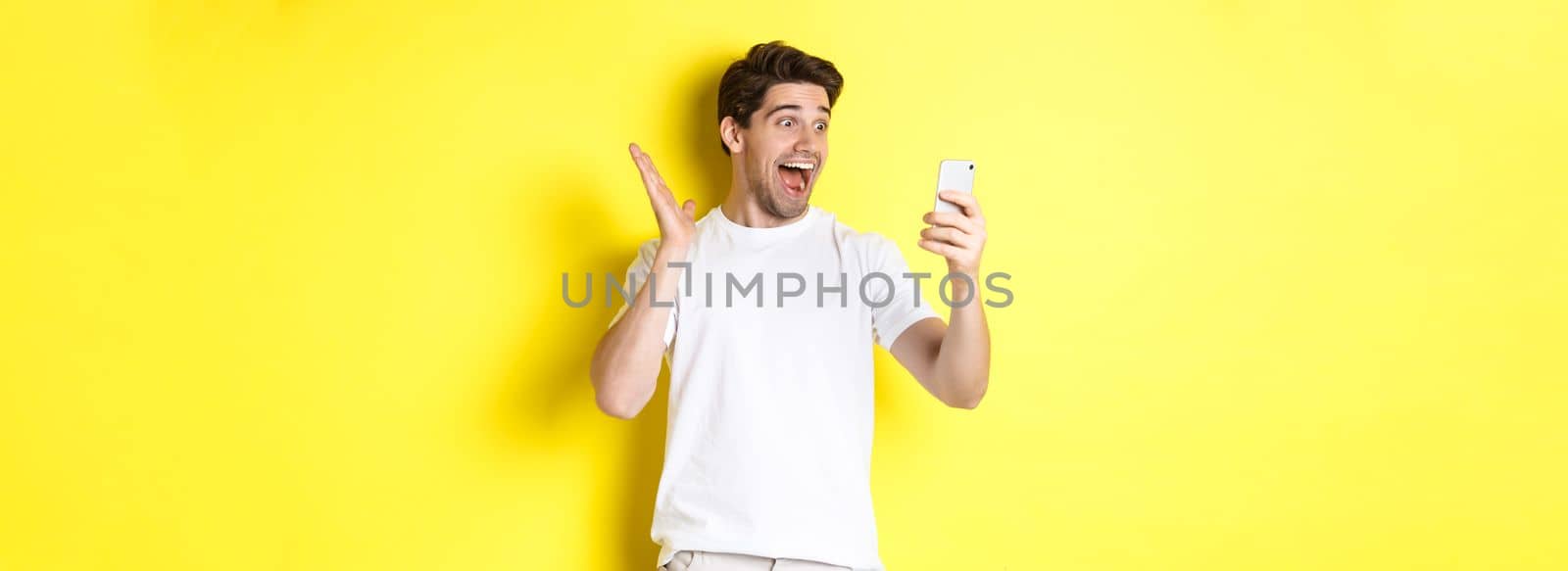 Surprised and happy man looking at mobile phone screen, reading fantastic news, standing over yellow background by Benzoix