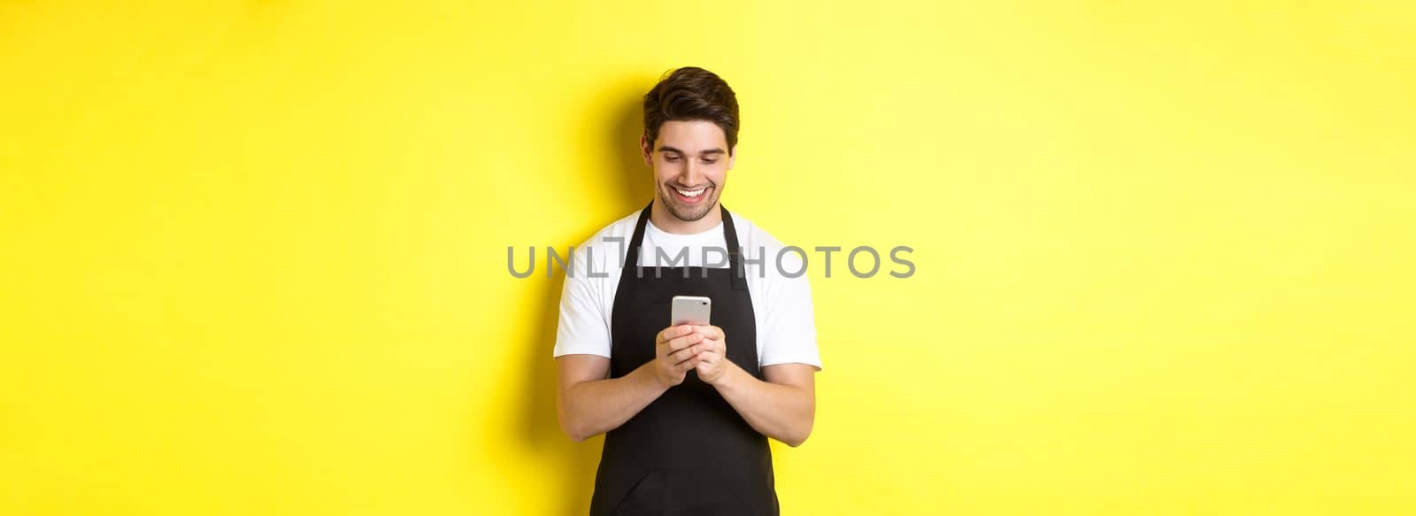 Waiter in black apron reading message on mobile phone, smiling happy, standing over yellow background by Benzoix