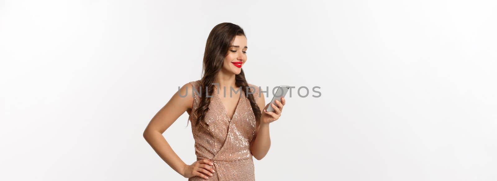 Christmas party and celebration concept. Happy young woman reading message on phone and smiling, using smartphone, wearing luxurious dress, white background.