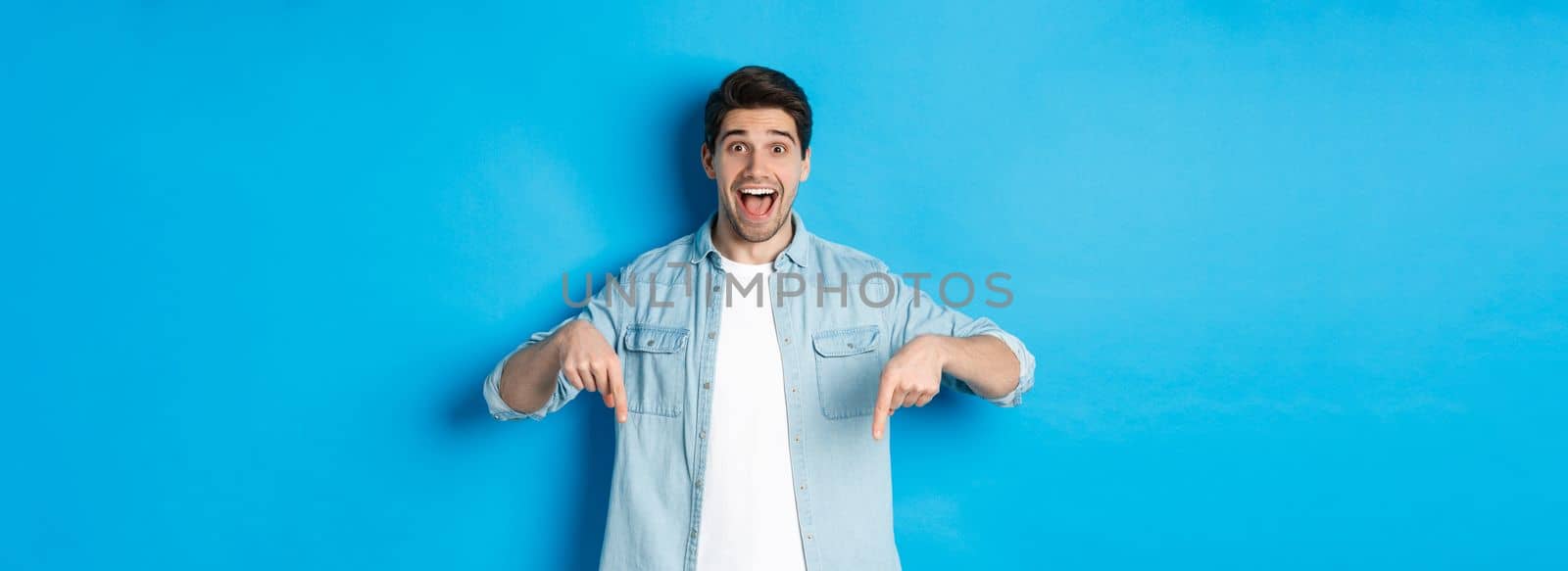 Image of excited handsome man pointing fingers down, making an announcement, standing against blue background.