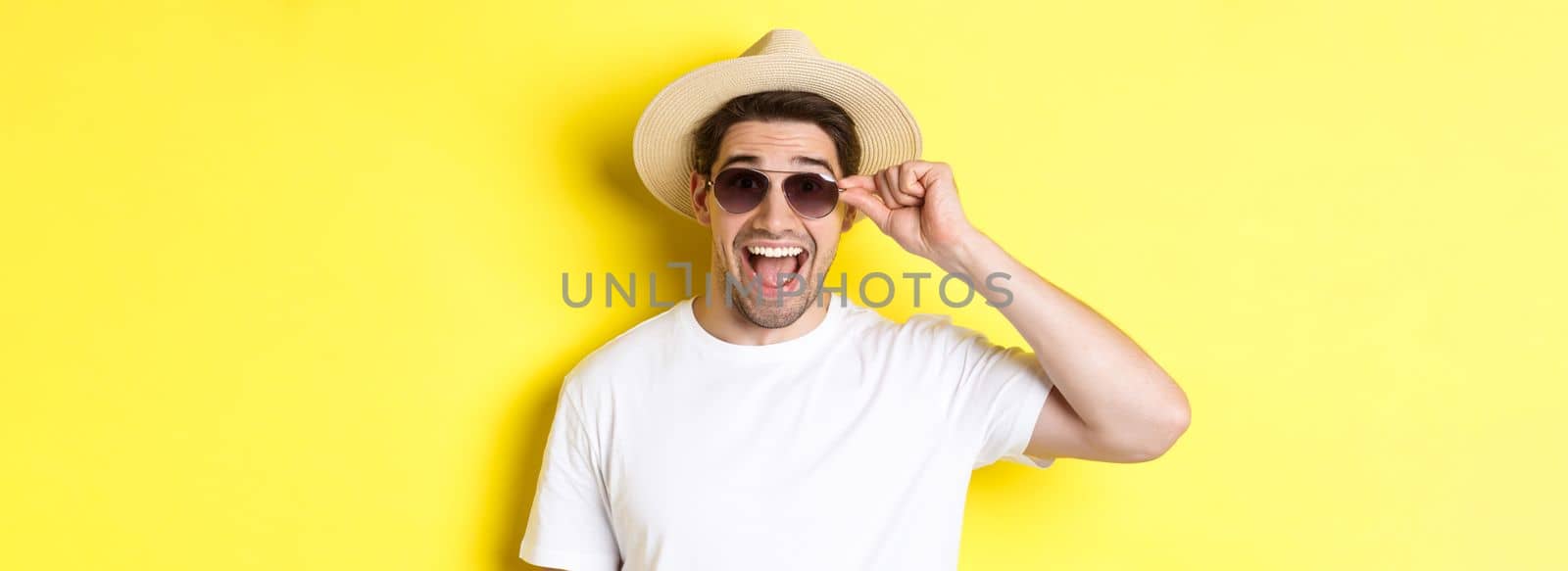 Concept of tourism and holidays. Close-up of happy man in summer hat and sunglasses enjoying vacation, standing over yellow background.