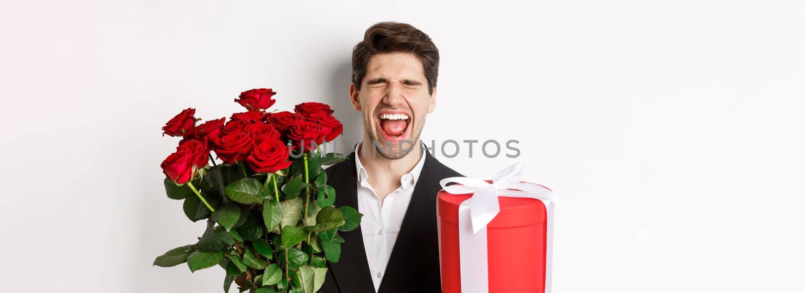 Image of sad guy in suit, got rejected and crying, holding bouquet of roses and present, standing miserable against white background.